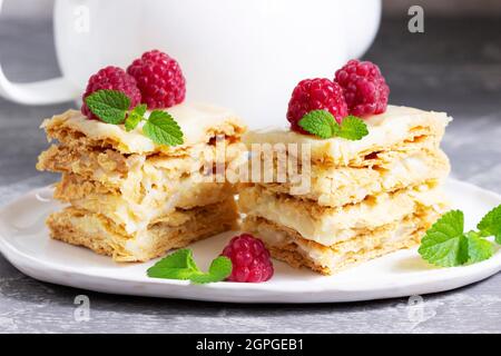 Pieces of Napoleon cake decorated with raspberries and lemon balm leaves on a light background. Stock Photo