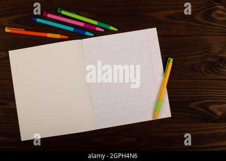 School notebook with oblique lines, multicolored pens on the table brown wooden background. Blank sheet of paper in notebook on the desk Stock Photo