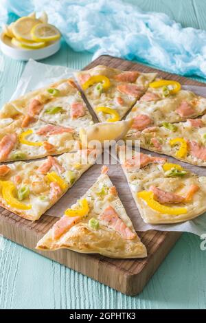 Pieces of tarte flambee with salmon, yellow pepper and spring onions on a wooden board on light green background, vertical Stock Photo