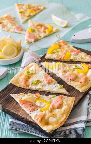 Pieces of tarte flambee with salmon, yellow paprika and spring onions on a wooden board on light green background, vertical Stock Photo