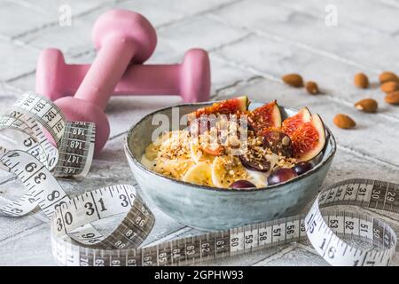 Bowl of yoghurt, fruits and nuts for a healthy diet breakfast or snack. Pink dumbbells and a tape measure on gray background, weight loss concept Stock Photo