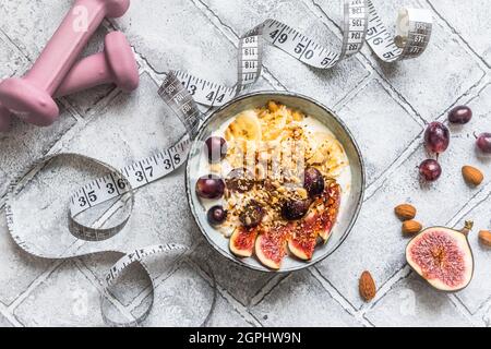 Bowl of yoghurt, fruits and nuts for a healthy diet breakfast or snack. Pink dumbbells and a tape measure on gray background, weight loss concept. Top Stock Photo