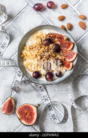 Bowl of yoghurt, fruits and nuts for a healthy diet breakfast or snack and a tape measure on gray background, weight loss concept. Top view. Stock Photo