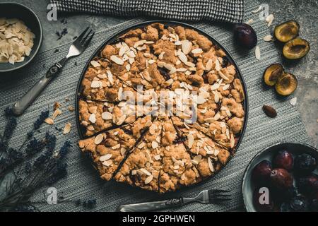 Whole plum cake with roasted almonds, cut into pieces on a gray plate on a gray background, top view Stock Photo