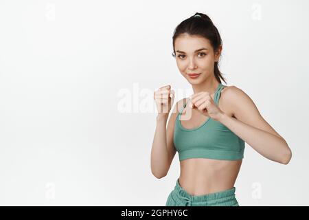 Image of fit and healthy fitness girl raising clenched fists, shadow boxing, doing workout. Young female athlete practice punching exercises, white Stock Photo