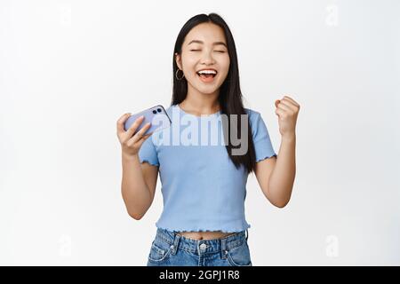 Enthusiastic smiling asian woman winning on mobile phone video game, looking satisfied and relieved, standing in t-shirt over white background Stock Photo