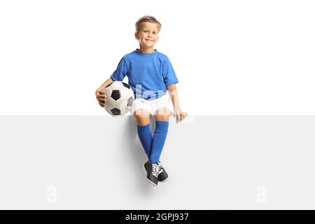 Cute little boy in a blue soccer jersey sitting on a blank panel and holding a ball isolated on white background Stock Photo