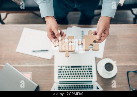 hand of businessman connecting jigsaw puzzle and laptop, coffee and document on the table. Business solutions, success and strategy concept Stock Photo