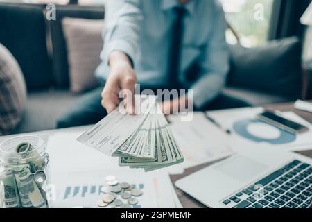 A Businessperson's hand giving cheque and dollar bill to customer and coin, laptop and graph chart on the desk at office. Payment by check, paycheck, Stock Photo