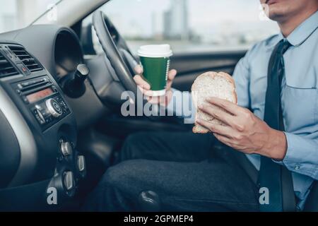 A businessman eating whole wheat toast and holding a coffee cup While driving on the road and he did not lose a seat belt. Stock Photo