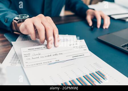 A Businessperson's hand giving cheque to customer and dollar bill, coin, laptop and graph chart on the desk at office. Payment by check, paycheck, pay Stock Photo