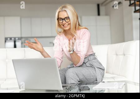 Smiling mature middle-aged blonde businesswoman in eyeglasses sitting on the couch in the living room, talking at the webcam of laptop, working remotely, communicating with colleagues or friends Stock Photo