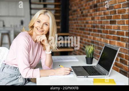 Positive charming mature blonde businesswoman sitting at the desk in front of laptop, working on a project. Freelancer enjoying remote work from home, looks at the camera and smiling Stock Photo