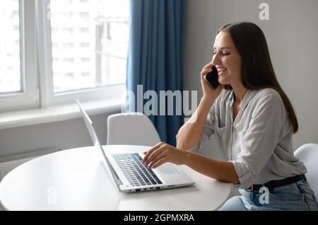 Work from home concept. Young female entrepreneur chatting on the smartphone sitting at the desk and using laptop, smiling woman has pleasant phone conversation during remote work Stock Photo