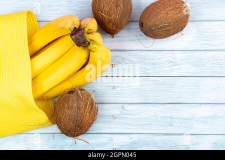 Whole coconuts lying in a yellow bag and bananas on a blue wooden background. Stock Photo