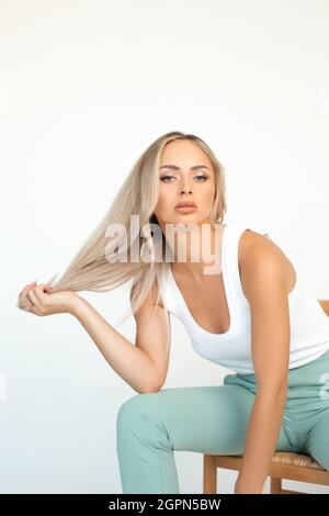 photo of young blonde girl in green pantsuit sitting on chair in studio on white background. Stock Photo