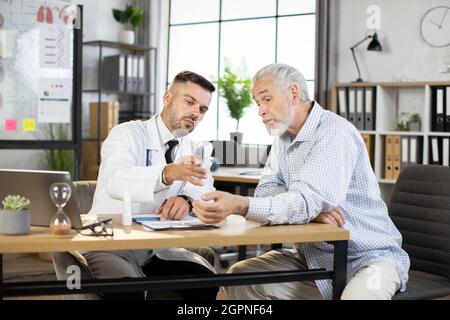 Professional male doctor using modern thermometer for measuring temperature of aged patient. Caucasian man visiting family doctor for regular checkup. Stock Photo