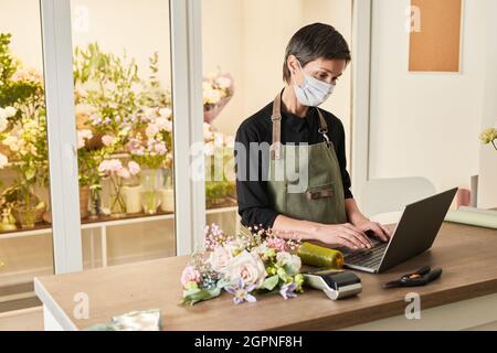 Portrait of female small business owner using laptop and wearing mask while managing flower shop, copy space Stock Photo