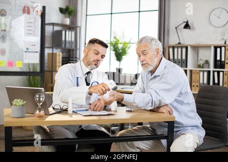 Caucasian medical worker in white lab coat measuring temperature of senior bearded man. Male doctor using modern thermometer during checkup of aged patient. Stock Photo