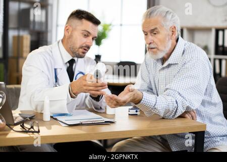 Medical practitioner in white lab coat using modern thermometer for examining health condition of aged patient. Caucasian senior man getting professional checkup at medical center. Stock Photo