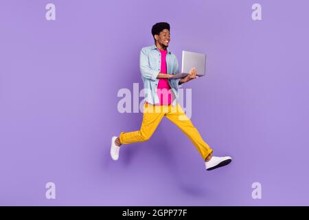 Profile photo of guy jump hold computer run fast wear blue shirt pants sneakers isolated purple color background Stock Photo