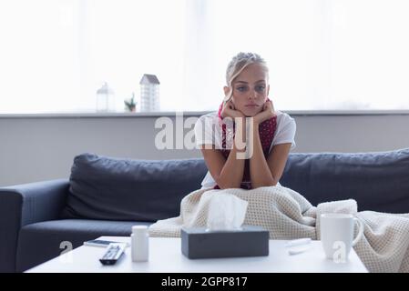 sad and sick woman in warm scarf sitting on sofa under blanket near paper napkins and pills container Stock Photo