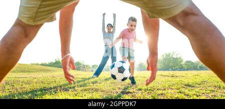 young happy family playing football outdoors into a public park garden. mom, dad and little child having fun together. cute kid kicking a ball Stock Photo