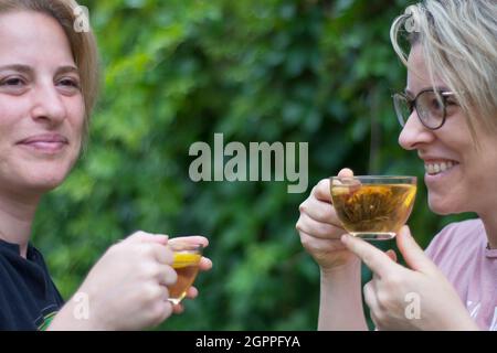 two young sister women are drinking blooming tea and chatting in the garden of their house. Selective Focus Glasses Stock Photo