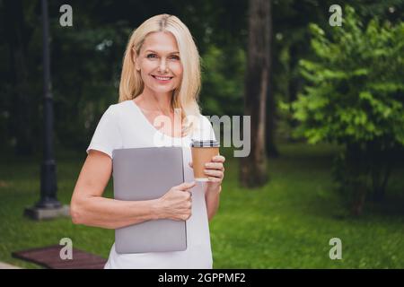 Photo of mature work blond lady hold laptop coffee wear white t-shirt in park alone Stock Photo