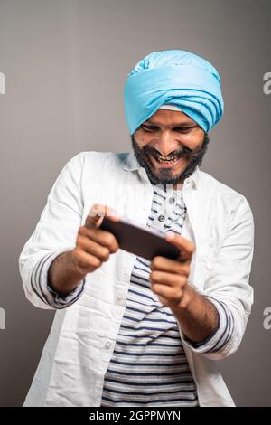 Young smiling sikh man busy playing video game on mobile phone - concept gaming addiction, using modern technology on studio background. Stock Photo