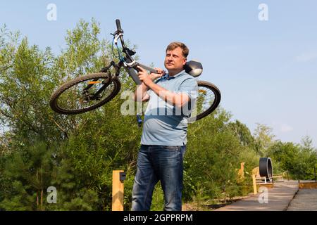 A frustrated man in the park carries a broken bicycle on his shoulder. Stock Photo