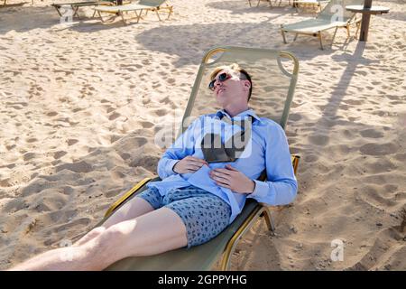 Young tired businessman lies on a chaise longue in a shirt with a tie and boxer shorts. Rest at sea, reboot. Downshifting Stock Photo