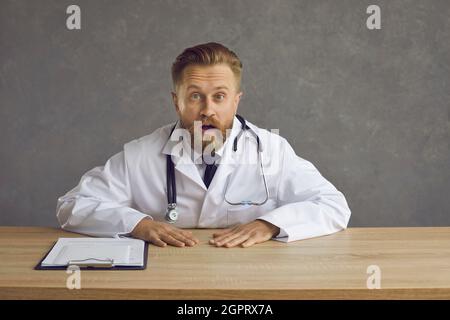 Young doctor sitting at desk and looking at camera with surprised face expression Stock Photo