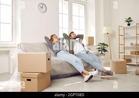 Happy couple taking a break and sitting on a couch while moving in their new house Stock Photo