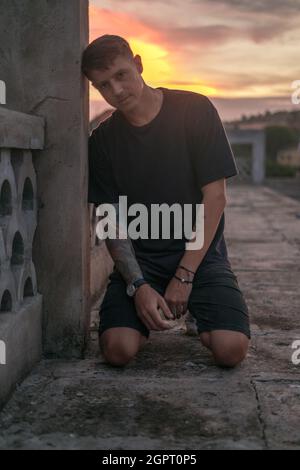 Portrait of young handsome Caucasian man with tattoo in black casual wearing, sitting on his knees and looking at camera. Photo on the sunset. Stock Photo