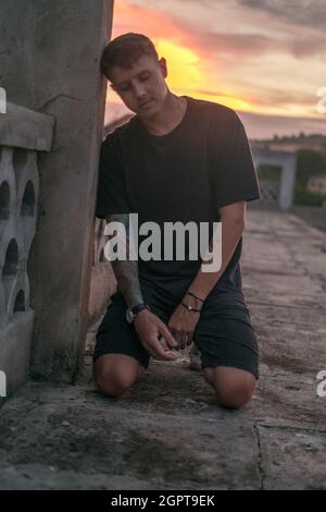 Portrait of young handsome Caucasian man with tattoo in black casual wearing, sitting on his knees and looking down. Photo on the sunset. Stock Photo