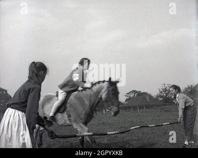 1950s, historical, outside in a field at at an equestrian event, a teenager girl rider on a horse about to jump over a wooden pole hend by two young  girls, West Sussex, England, UK. Riders on horses jumping over obstacles or barriers, sometimes hand-held as seen here, are a common sight at horse shows, with show jumping being part of a group of English riding equestrian events. Stock Photo