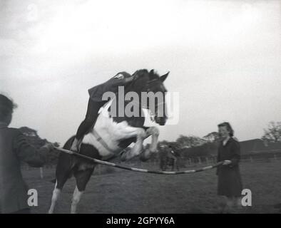 1950s, historical, outside in a field at at an amateur equestrian event, a rider on a horse jumping over a wooden pole held by two girls standing opposite each other, West Sussex, England, UK. Jumping over obstacles or barriers, sometimes hand-held as seen here, are a common sight at horse shows, with show jumping being part of a group of English riding equestrian events. Stock Photo