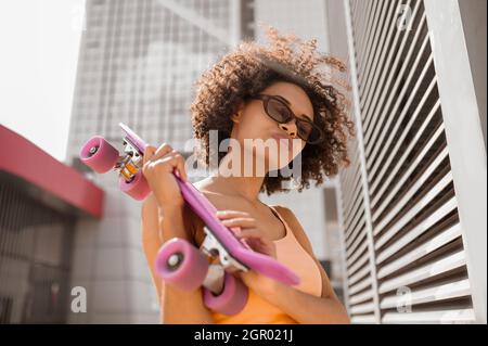 A young mulatta with a skateboard in hands standing near the wall Stock Photo
