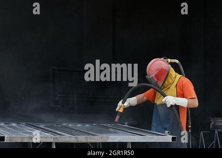 Industrial worker dressed in protective helmet and clothes sandblasting metal details for removing dust and rust. Young man cleaning surface of steel Stock Photo