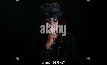 Creepy senior woman with Halloween witch makeup presses index finger to lips makes silence gesture Stock Photo