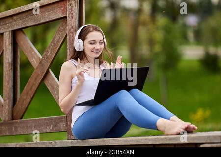 Smiling woman in cordless headphones works with laptop online in park. Stock Photo