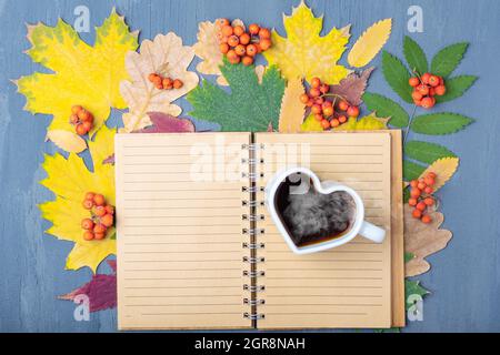 White mug in the shape of a heart with steaming coffee or tea and a blank lined craft notepad on a background of autumn fallen dry colorful leaves, to Stock Photo