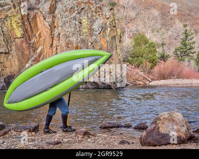 kayaker is carrying a whitewater inflatable kayak on a shore of mountain river in early spring - Poudre River in northern Colorado Stock Photo