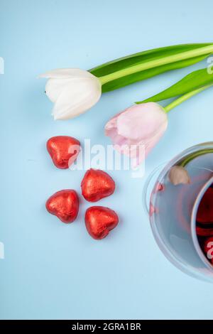 top view of white and pink color tulips with a glass of wine and heart shaped chocolate candies in red foil scattered on blue background