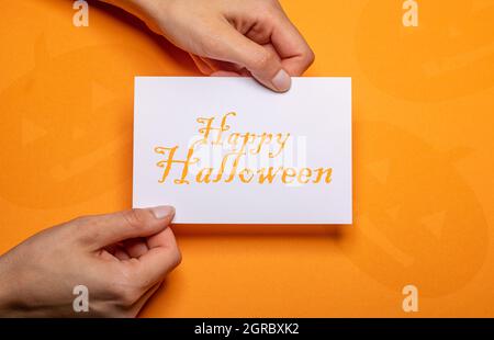 female hands holding a piece of paper with the inscription happy halloween in orange writing over orange background cardboard Stock Photo