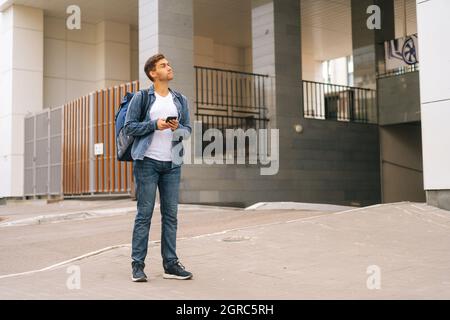 Front view of confused handsome male courier with thermo backpack using navigation app on phone standing in city street. Stock Photo