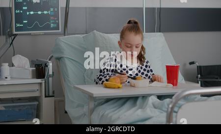 Portrait of hospitalized little child resting in bed eating healthy food meal during recovery examination after suffering medical surgery. Sick kid wearing oxygen nasal tube having breakfast nutrition Stock Photo