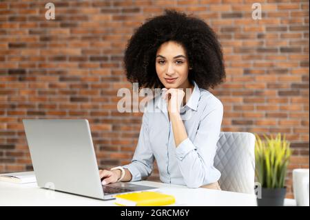 Smiling intelligent biracial woman with Afro hairstyle using laptop for work online sitting at the desk and looks at camera in the coworking space, female freelancer typing on the keyboard Stock Photo
