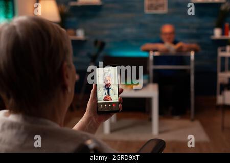 Elder woman with disability using video call for telemedicine at home. Aged adult talking to doctor for checkup on online remote conference with smartphone sitting in wheelchair Stock Photo